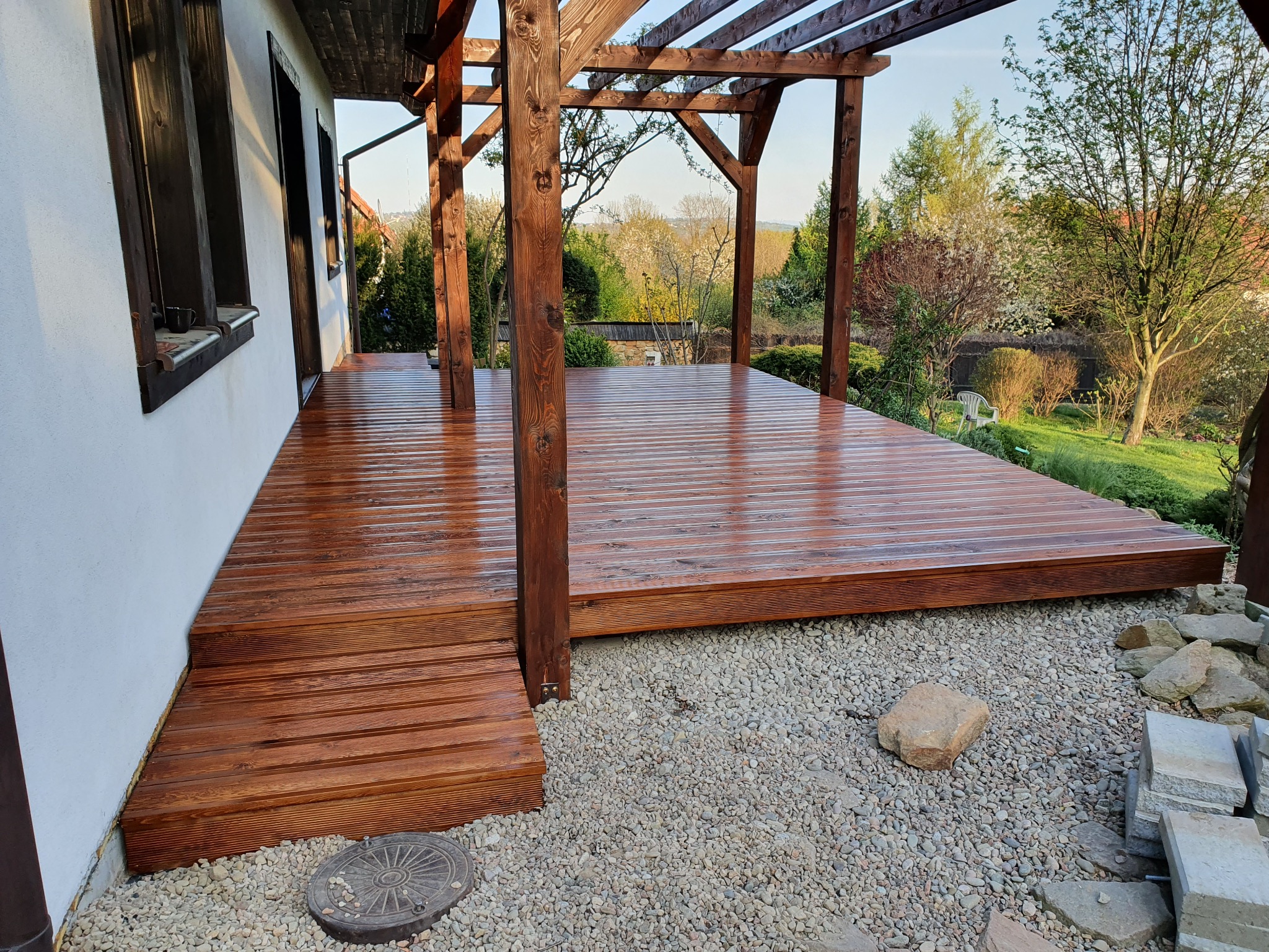 Świeżo zaimpregnowany taras z drewna egzotycznego, zadaszony drewnianą pergolą, otoczony żwirowym podłożem i zielenią ogrodu.
