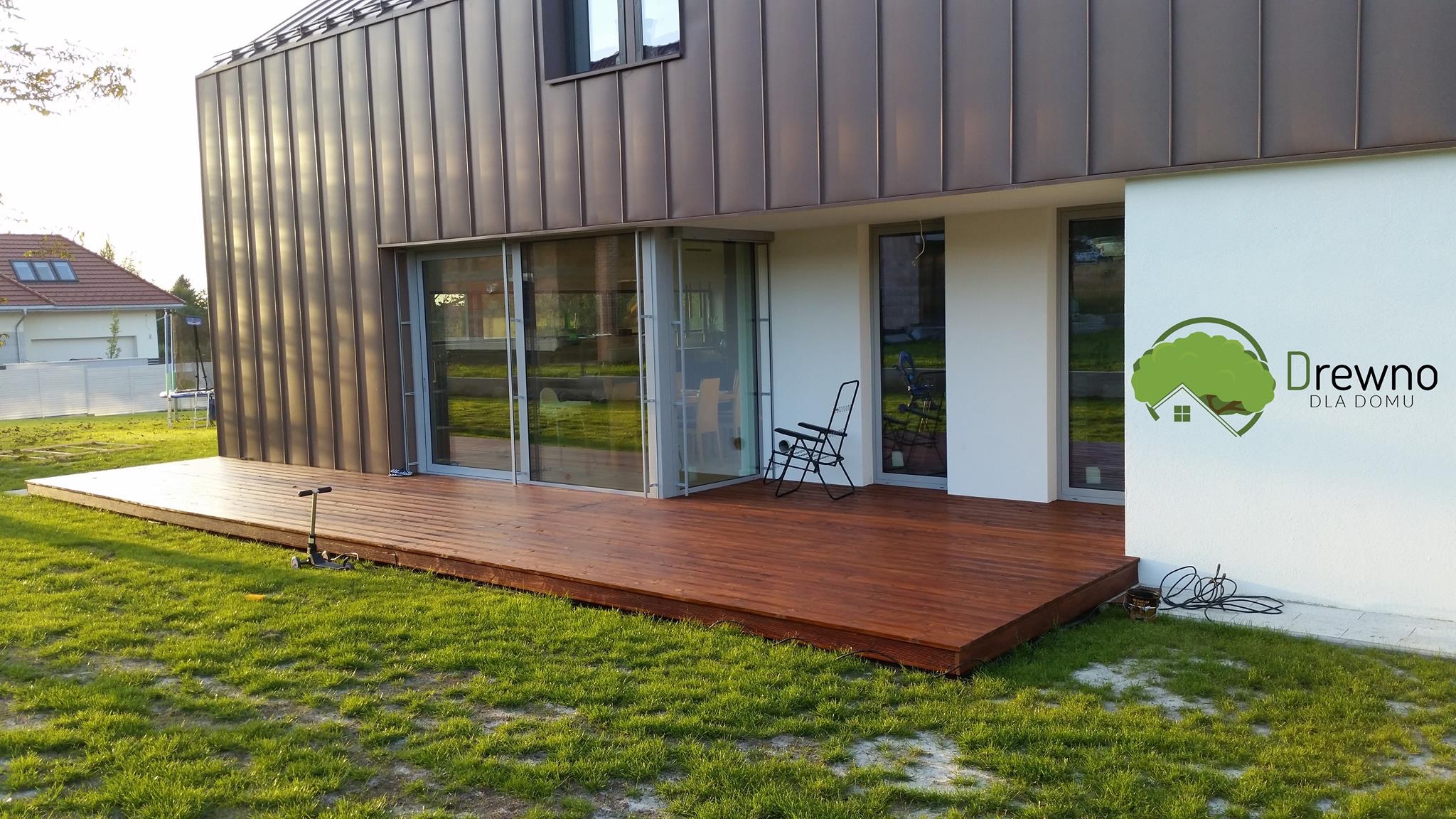 Nowoczesny dom z drewnianym tarasem, na trawniku stoi hulajnoga, na ścianie logo firmy 'Drewno dla domu'.