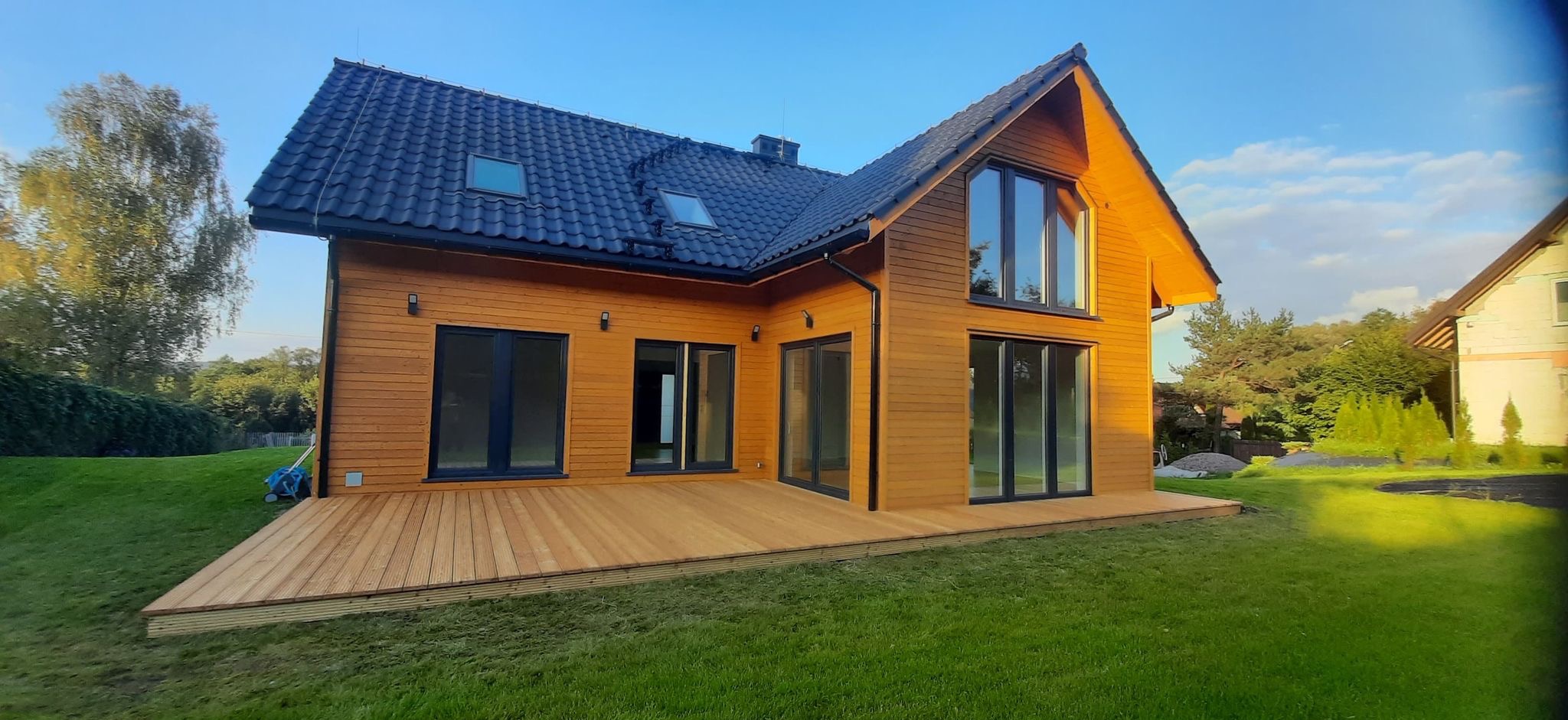Nowoczesny dom z drewna z dużym tarasem z desek, czarne okna i dachówka, zielony trawnik, błękitne niebo.