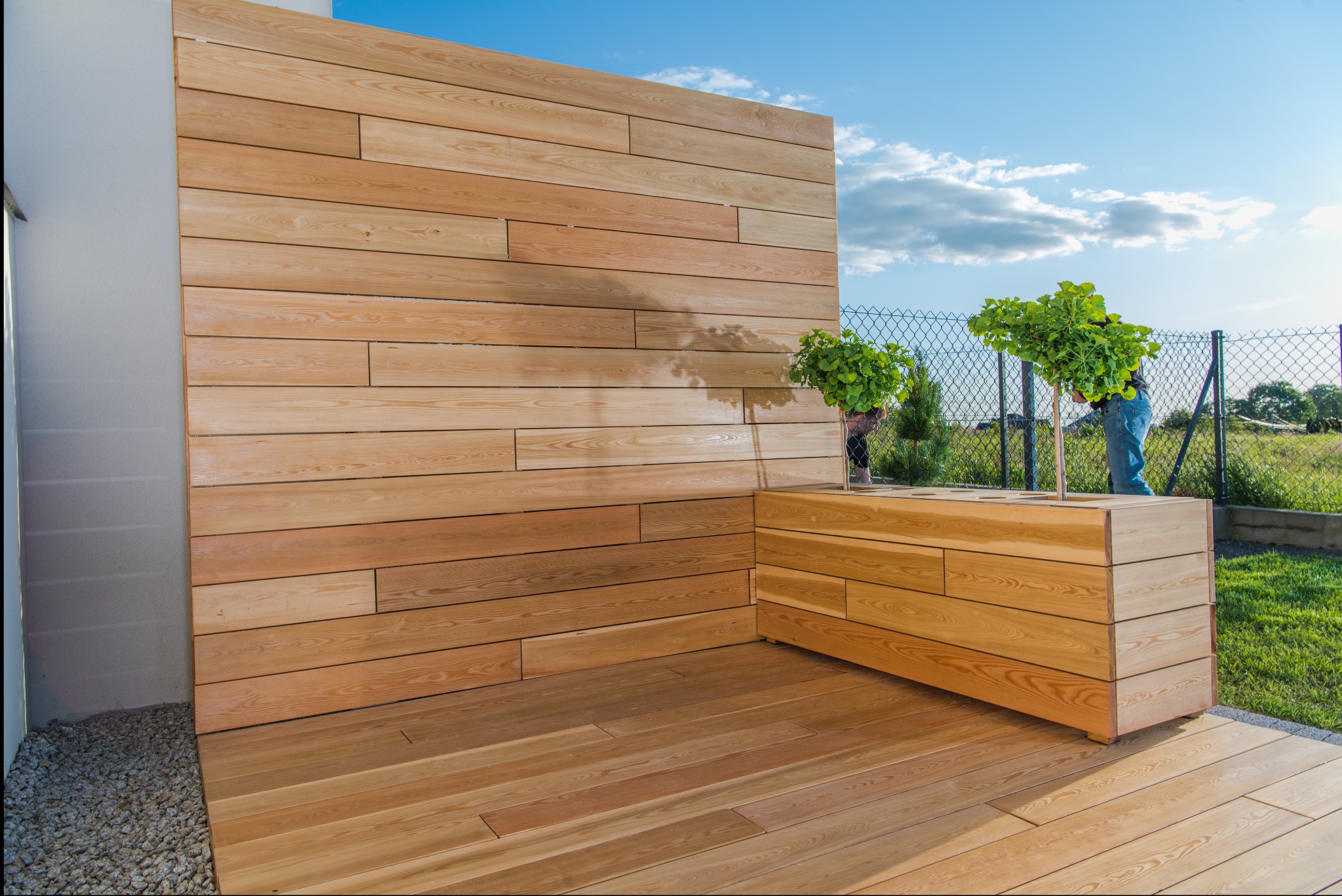 Nowoczesny taras z jasnego drewna z drewnianą ścianką i donicami, widoczne osoby pielęgnujące rośliny, błękitne niebo w tle.