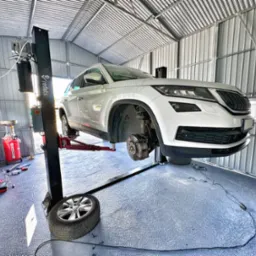 Biała Skoda Kodiaq na podnośniku w blaszanym garażu, brak koła, widoczne narzędzia i zbiornik na olej, oświetlenie dzienne z okna.