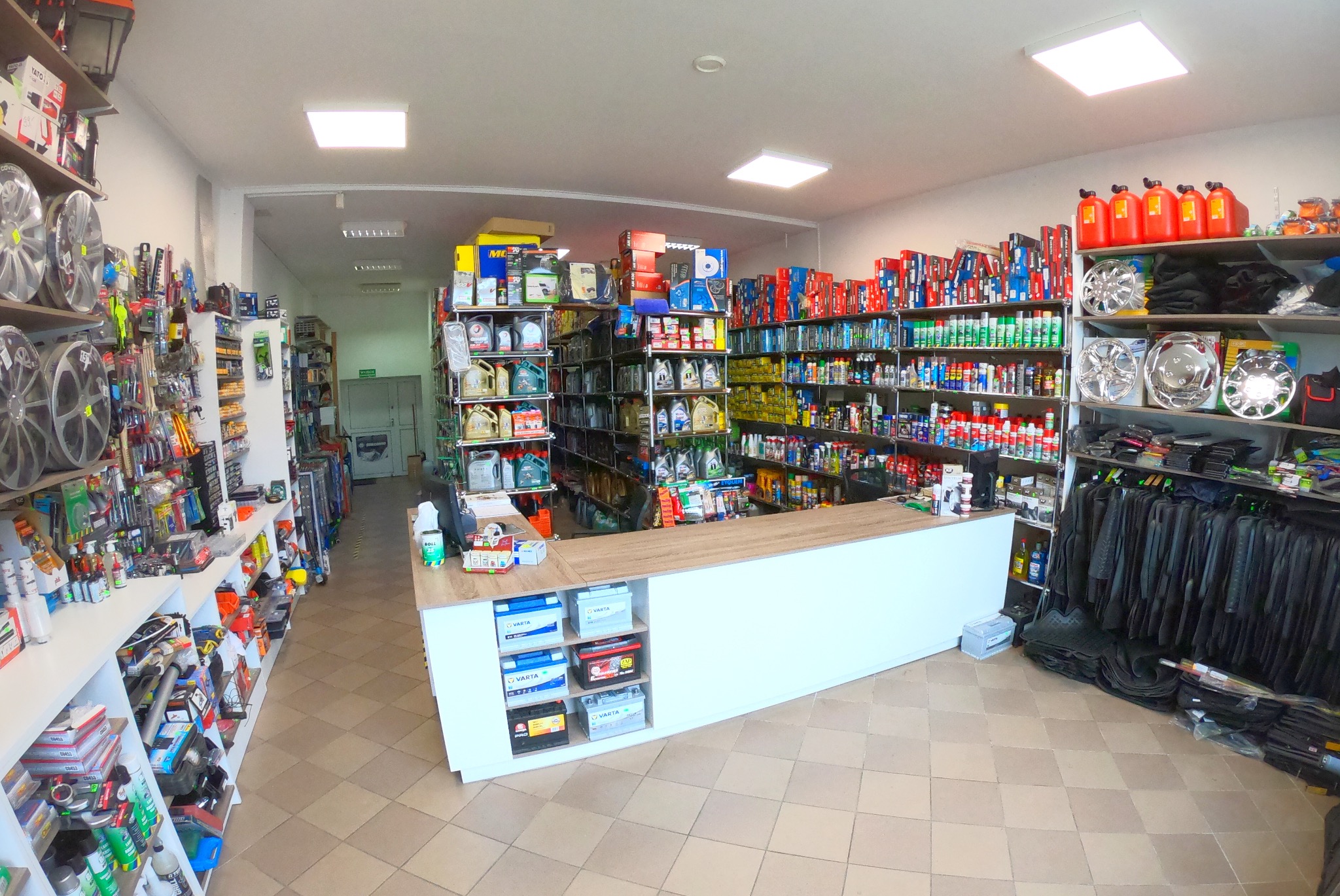 Wnętrze sklepu z akcesoriami i częściami samochodowymi: regały wypełnione olejami, płynami, narzędziami i felgami, widoczna lada sklepowa z akumulatorami.