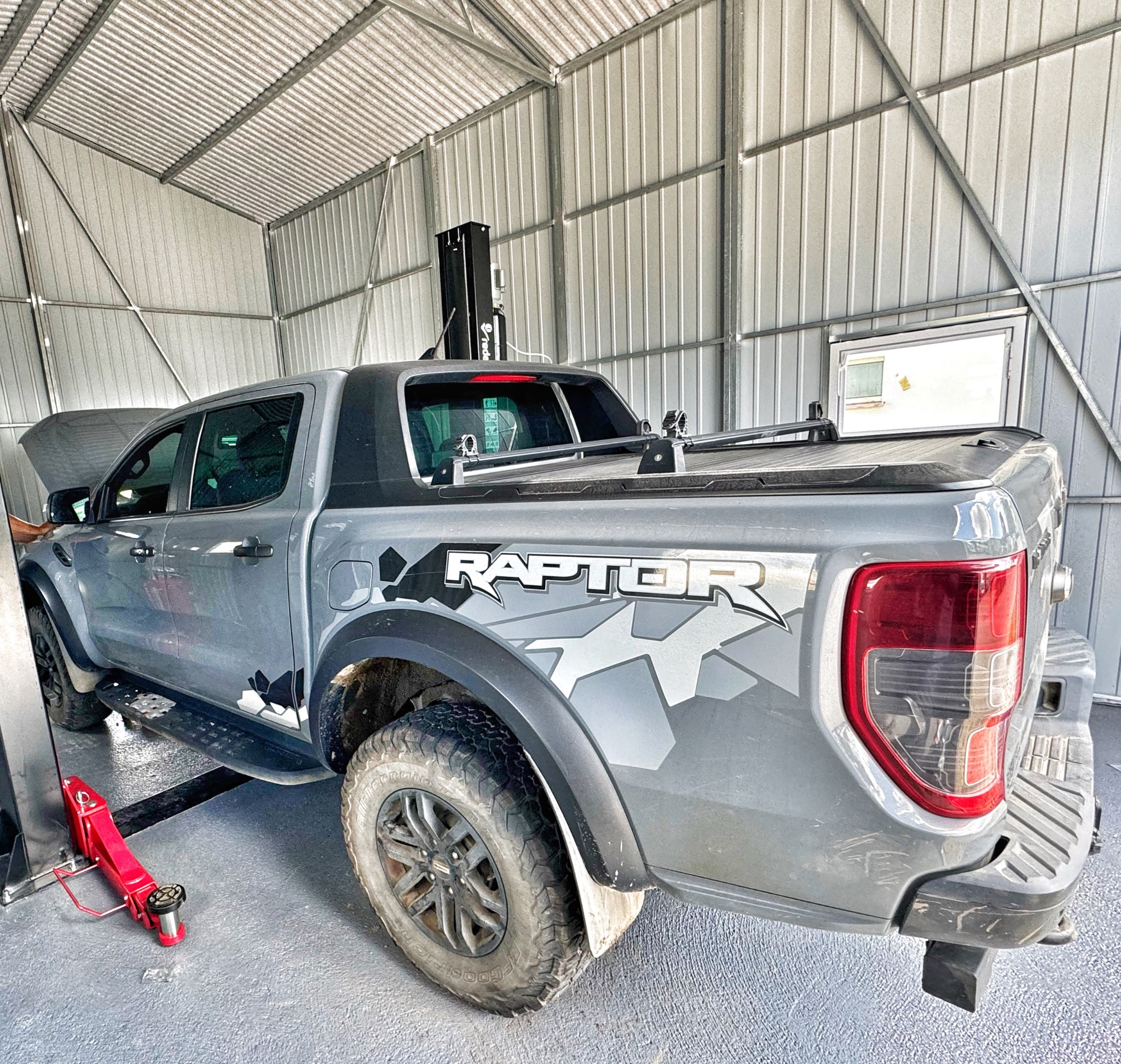 Szary Ford Ranger Raptor na podnośniku w warsztacie z otwartą maską, widoczny czerwony podnośnik hydrauliczny i stalowa konstrukcja budynku.
