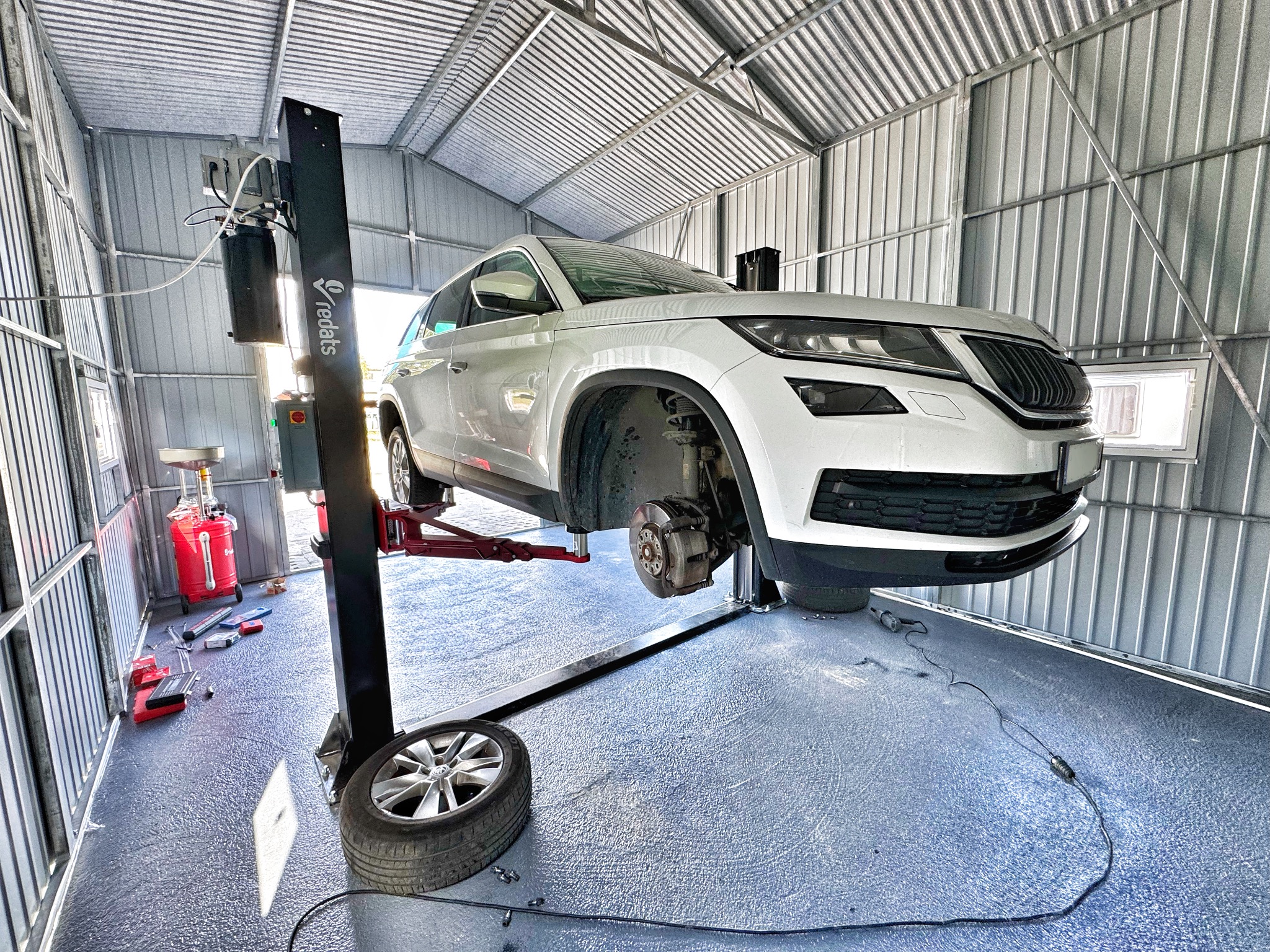 Biała Skoda Kodiaq na podnośniku w blaszanym garażu, brak koła, widoczne narzędzia i zbiornik na olej, oświetlenie dzienne z okna.