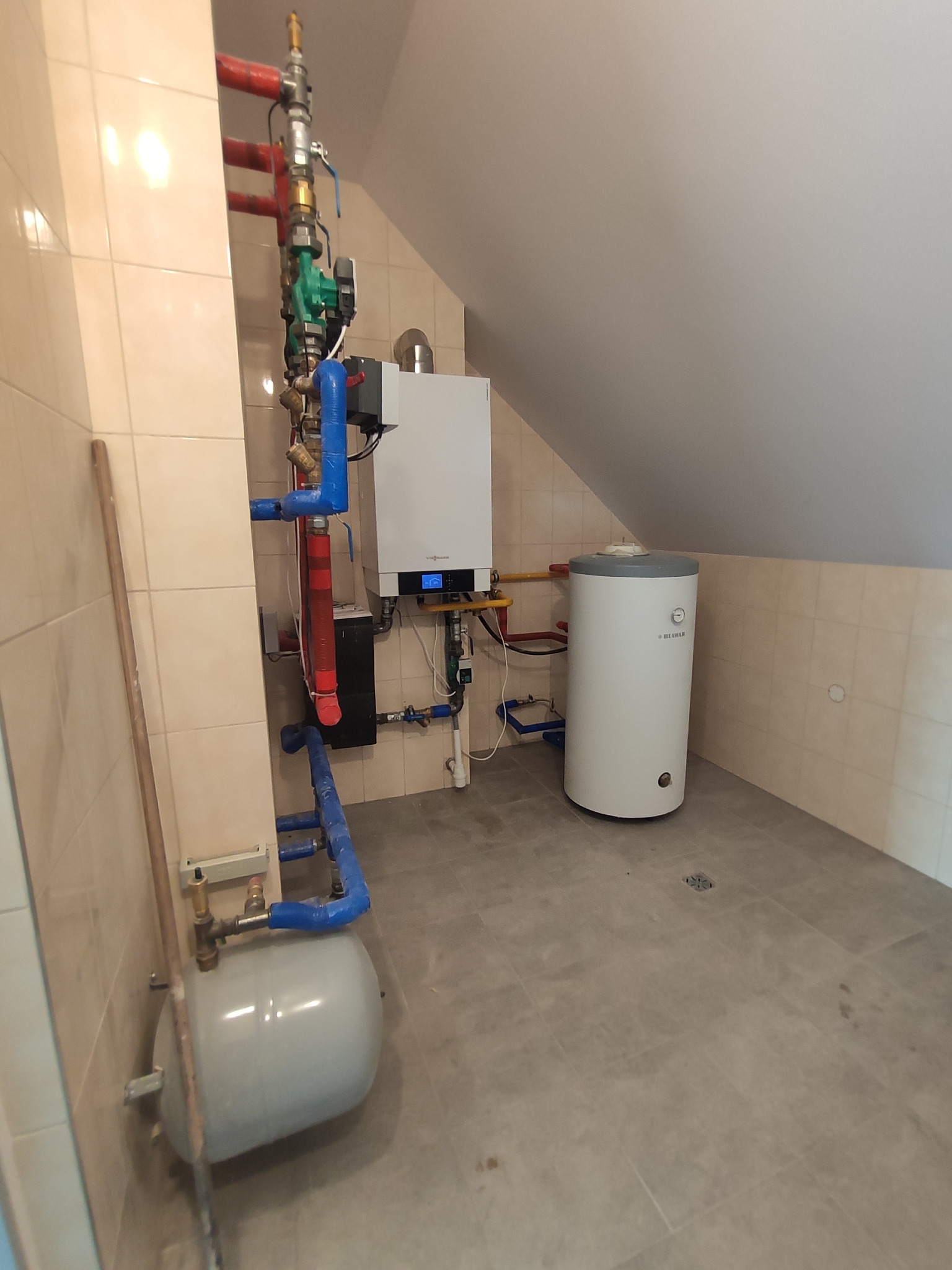 Nowoczesny piec centralnego ogrzewania Viessmann z podłączonym zasobnikiem ciepłej wody użytkowej, zainstalowany w pomieszczeniu ze skośnym sufitem i wyłożonym płytkami, widoczne rury instalacji...