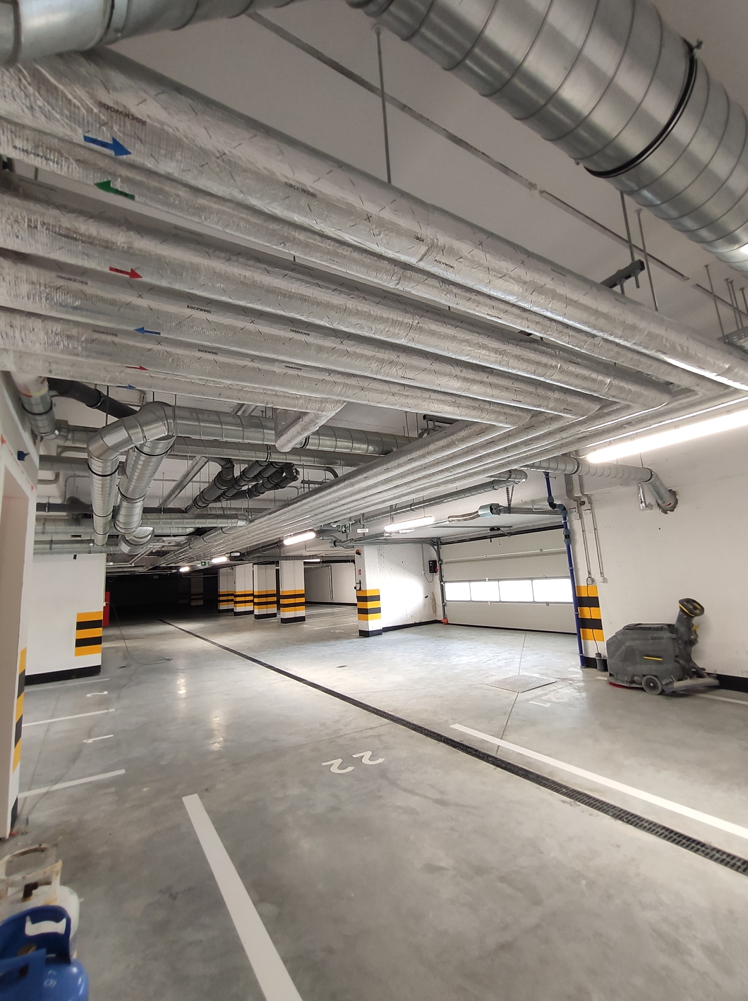 Wnętrze parkingu podziemnego z widocznymi licznymi, izolowanymi rurami wentylacyjnymi pod sufitem, oznaczonymi strzałkami w różnych kolorach, oraz maszyną czyszczącą podłogi w tle.