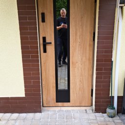 BAMAR MARCIN SIDOR - Drewniane drzwi zewnętrzne z czarnym, wąskim przeszkleniem, zamontowane w elewacji domu z ceglastą i żółtą ścianą. W szybie odbija się osoba.