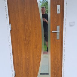 BAMAR MARCIN SIDOR - Brązowe drzwi wejściowe z ozdobnym przeszkleniem i srebrnymi elementami, widoczne ślady montażu na ścianie, odbicie osoby w przeszkleniu.