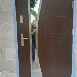 Nowoczesne drzwi zewnętrzne z przeszkleniem i brązowym wykończeniem, zamontowane w surowym murze z widocznym oknem nadprożowym. W lustrze odbija się osoba.