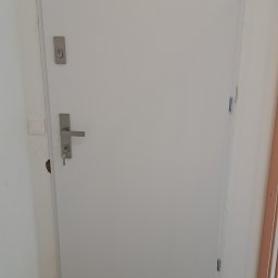 BAMAR MARCIN SIDOR - Białe drzwi z zamkiem i kluczem w korytarzu z czarno-białą podłogą w szachownicę. Widoczne zawiasy i listwa przypodłogowa.