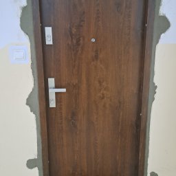 BAMAR MARCIN SIDOR - Ciemnobrązowe drzwi z wizjerem i srebrną klamką, osadzone w ościeżnicy z widocznymi śladami świeżego montażu i zaprawy. Wykończenie w trakcie prac, widoczna wycieraczka.