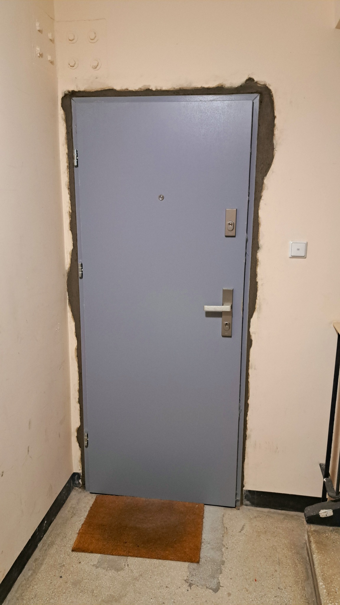Szare drzwi wejściowe w trakcie montażu, widoczne ślady zaprawy wokół ościeżnicy, brązowa wycieraczka przed drzwiami.