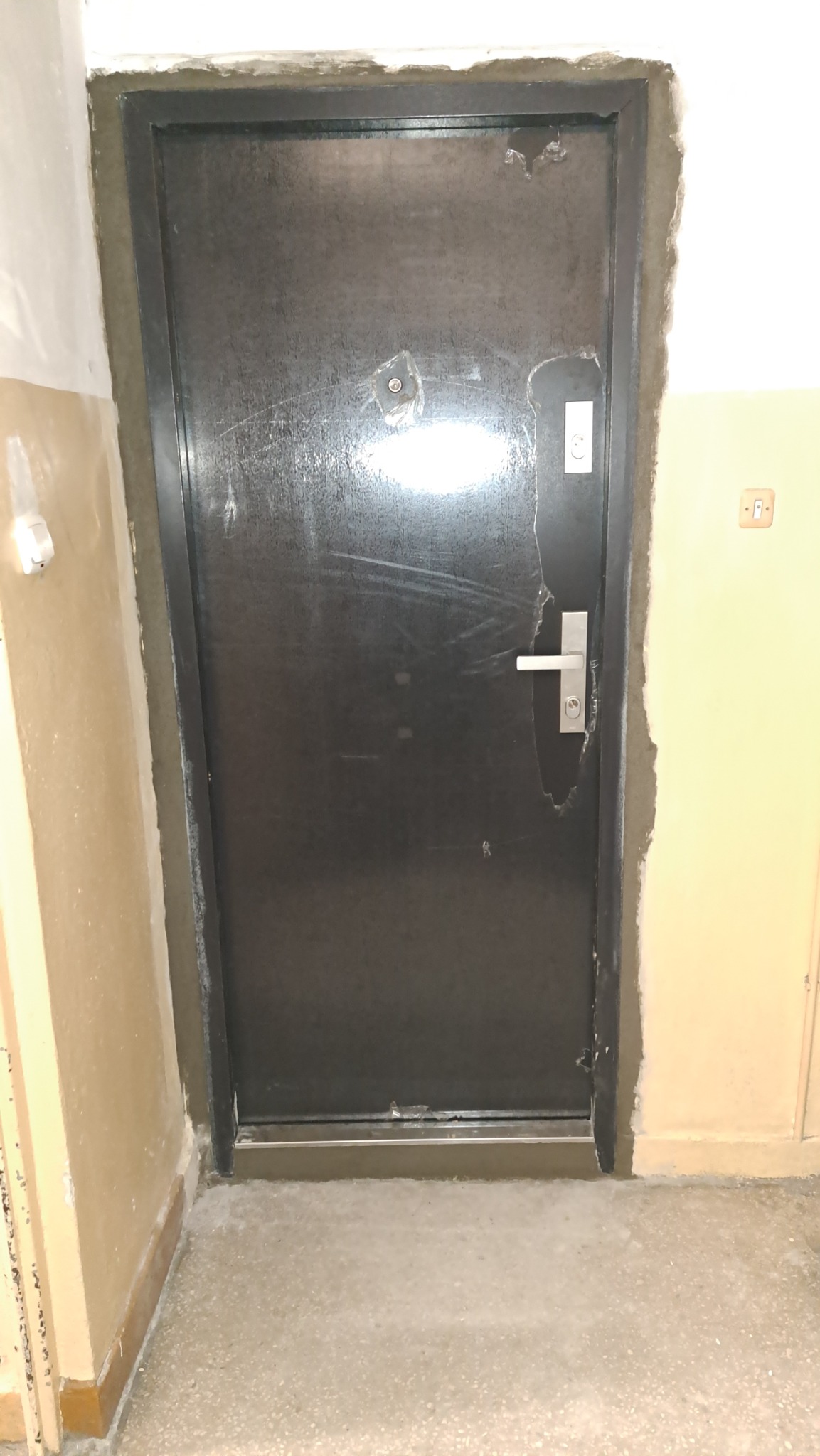 Ciemne drzwi wejściowe w trakcie montażu, widoczne świeże obramowanie cementowe, surowe wykończenie wnętrza.