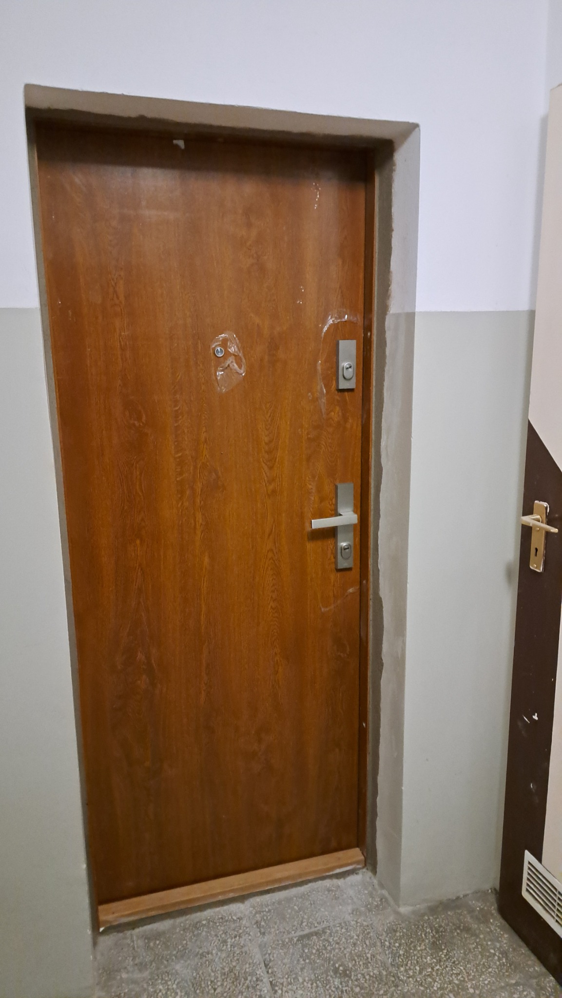 Drewniane drzwi wejściowe z metalowym zamkiem i klamką, zabezpieczone folią ochronną na nowym osiedlu, widoczne ślady po remoncie.