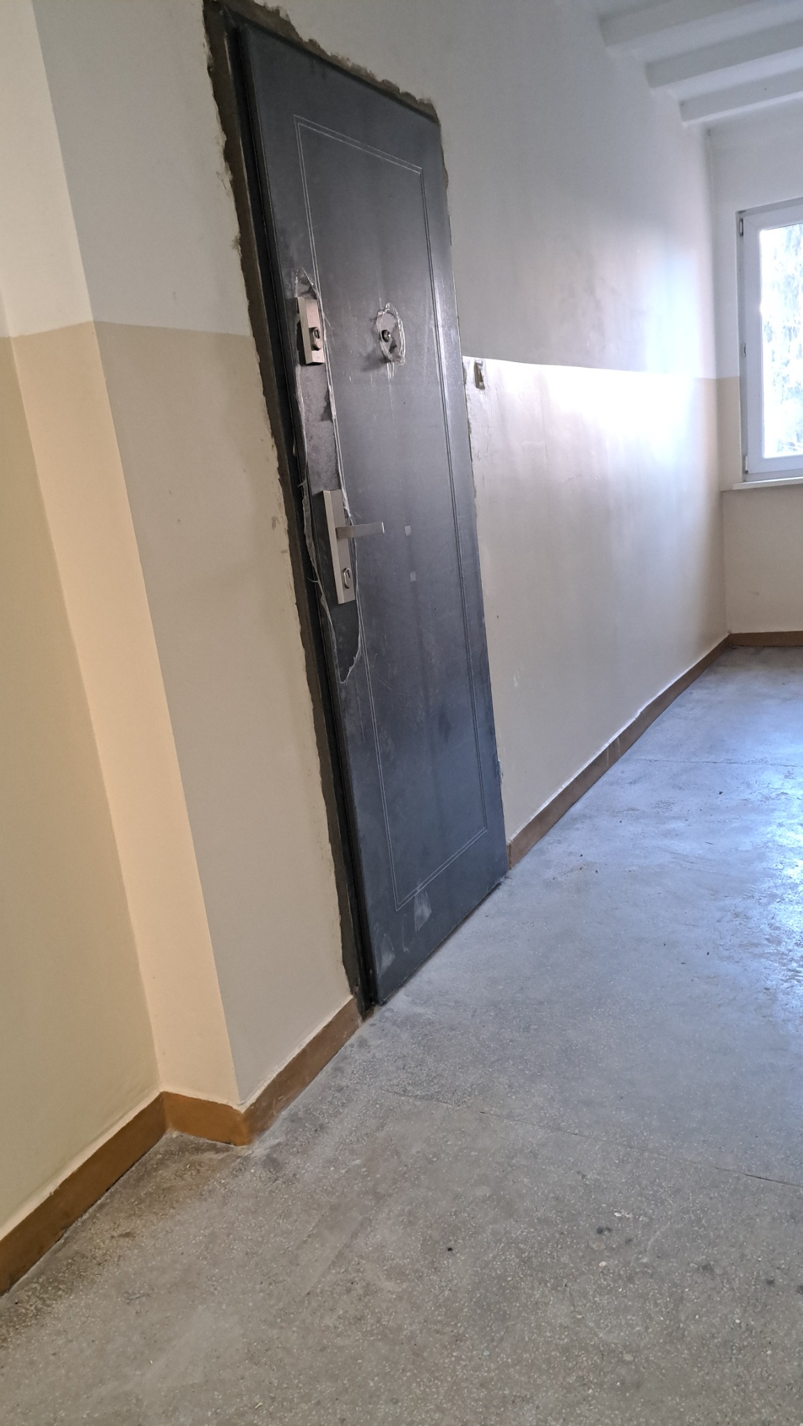 Ciemne drzwi wejściowe w trakcie montażu, widoczne ślady po pianie montażowej i niedokończone wykończenie ściany w korytarzu bloku.