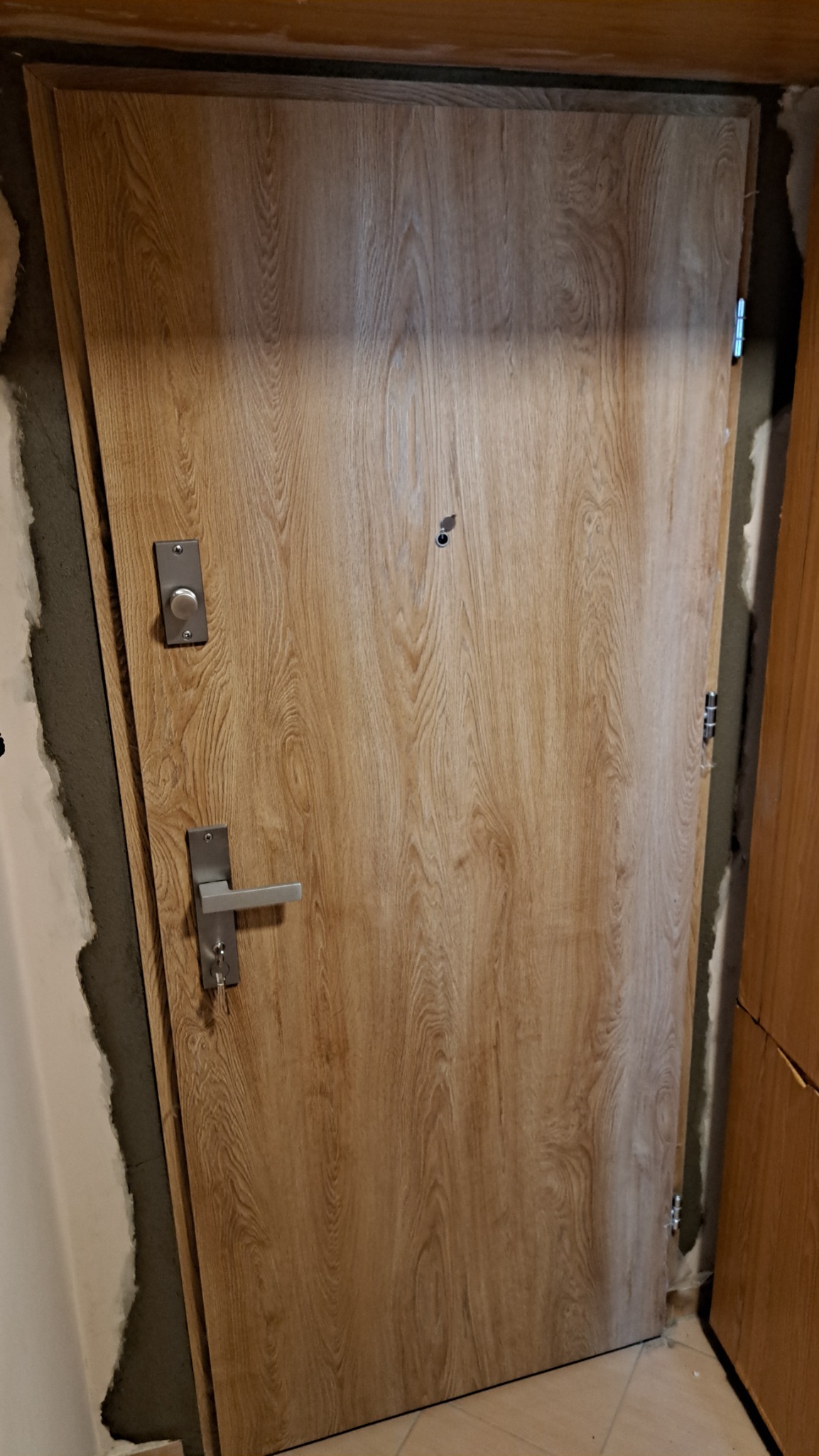 Jasne drzwi zewnętrzne z wizjerem i srebrną klamką, zamontowane w surowym otworze, z widocznymi śladami montażu i fragmentem podłogi.