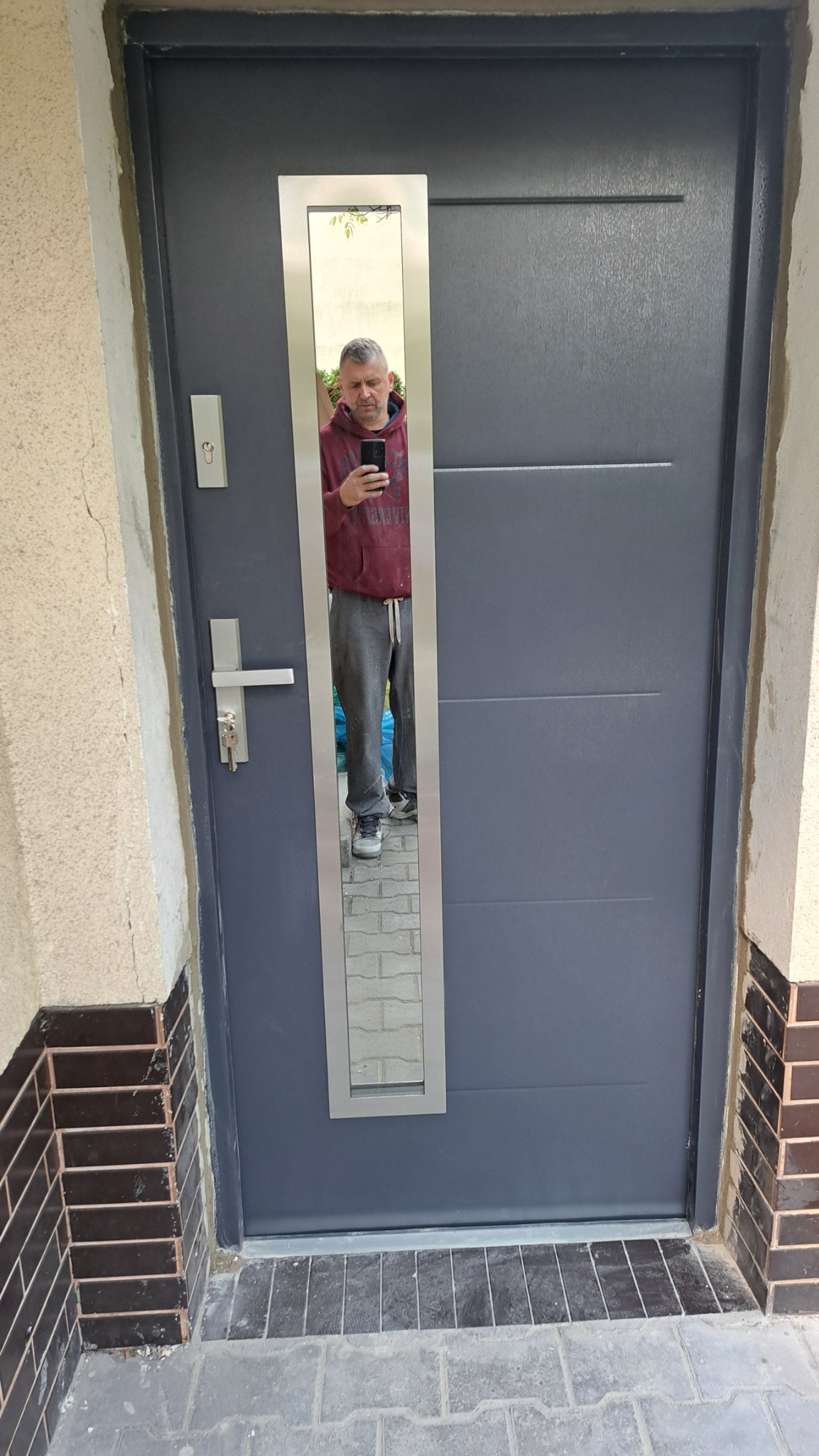 Szare drzwi zewnętrzne z podłużnym, lustrzanym wizjerem, w którym odbija się mężczyzna robiący zdjęcie telefonem. Nowe drzwi zamontowane w wejściu do budynku.