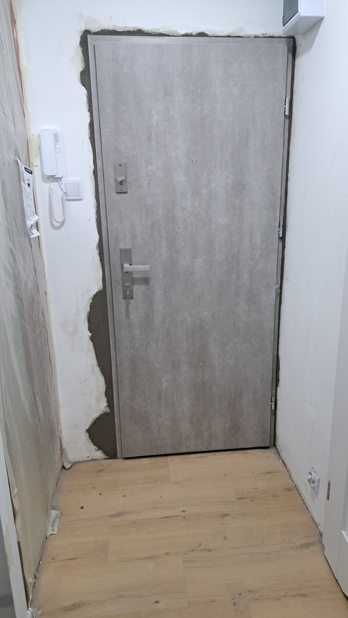 Szare drzwi z zamkiem i kluczami w surowym otworze, widoczne ślady montażu na ścianie. Podłoga wyłożona panelami.