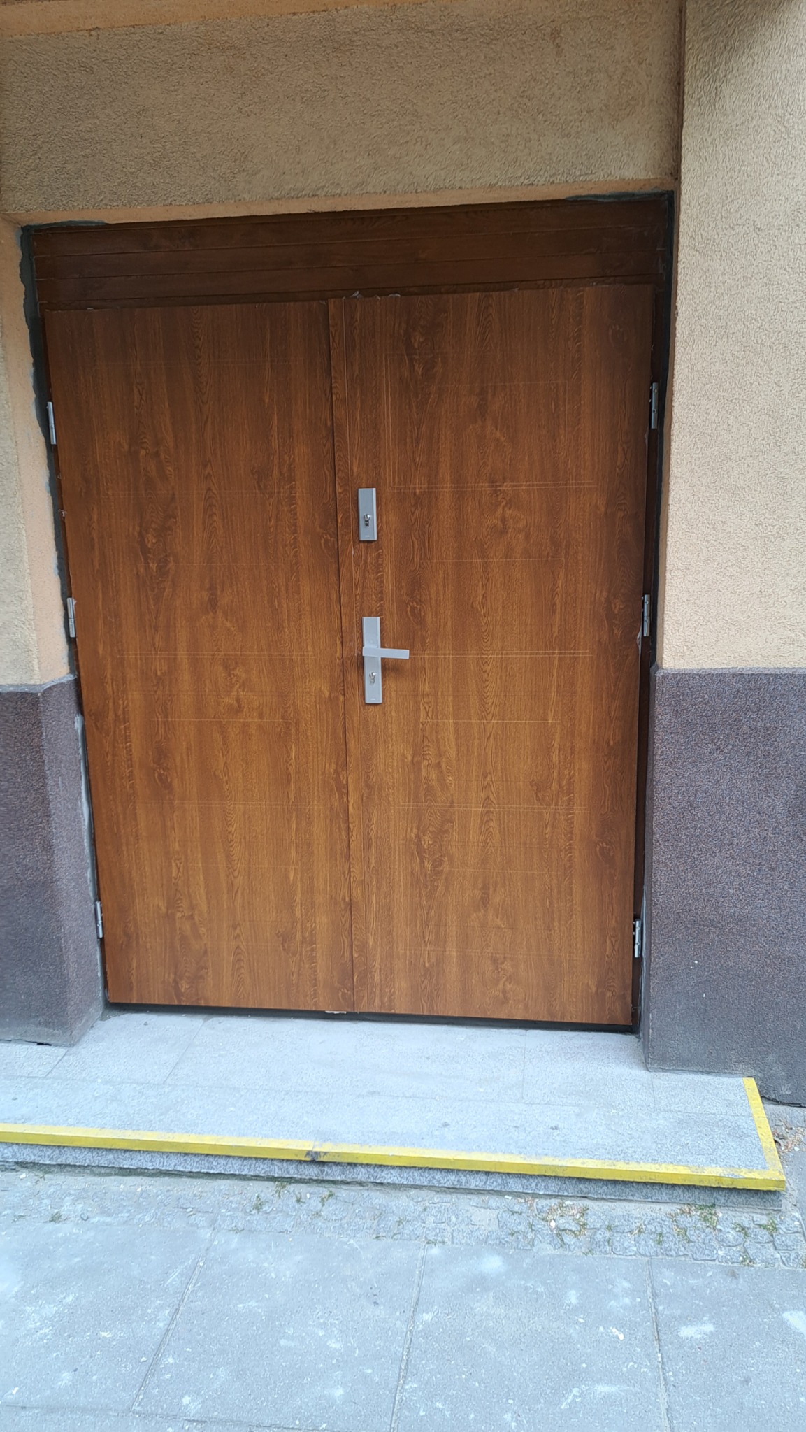 Brązowe, dwuskrzydłowe drzwi zewnętrzne z metalową klamką i zamkiem, zamontowane w beżowej elewacji budynku z ciemnoszarą podmurówką i żółtym progiem.