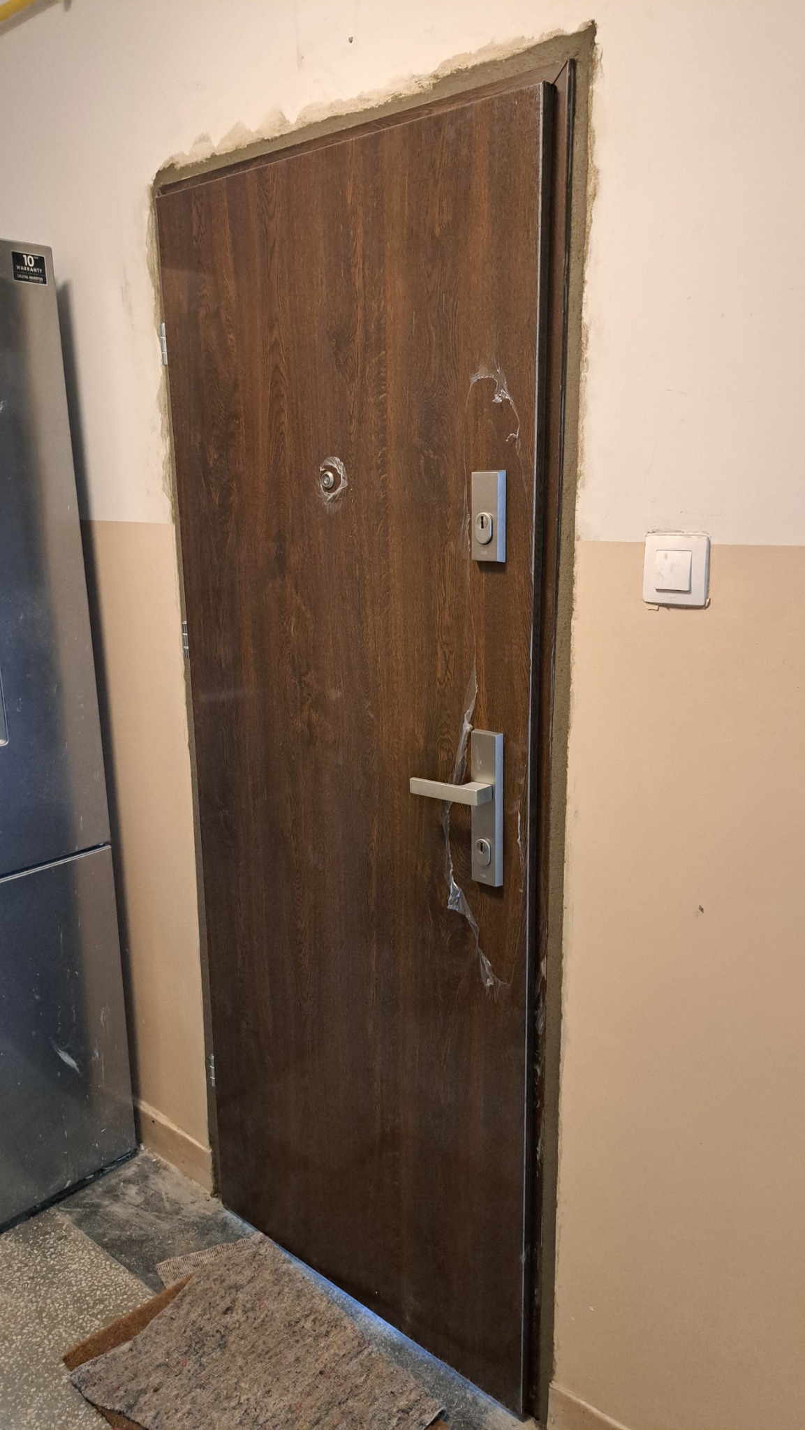 Nowe, brązowe drzwi z wizjerem i srebrną klamką, jeszcze z folią ochronną, zamontowane w ościeżnicy. Widoczny fragment lodówki.