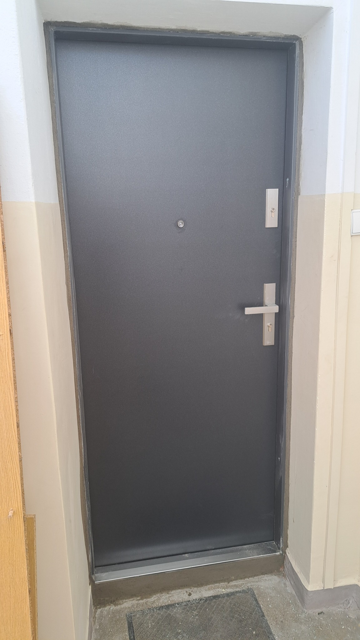 Szare drzwi zewnętrzne z wizjerem i srebrną klamką, osadzone w beżowej futrynie, widoczne na tle szarej podłogi z elementami kratki.