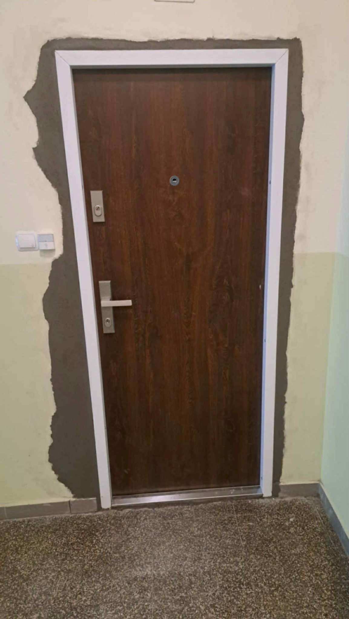 Montaż brązowych drzwi zewnętrznych z wizjerem i srebrną klamką. Widoczne ślady po montażu, szpachla na ścianie. Podłoga z lastryko.