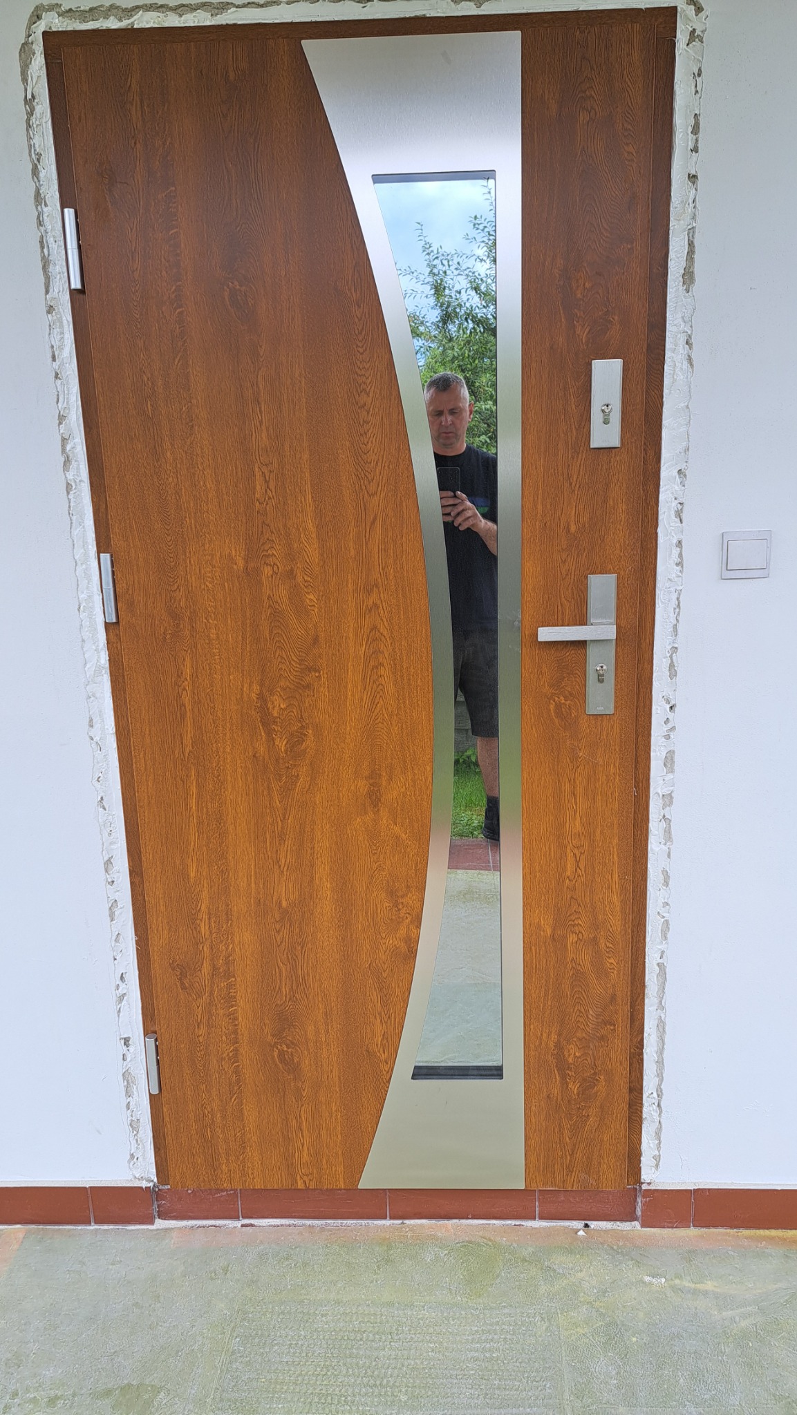 Brązowe drzwi wejściowe z ozdobnym przeszkleniem i srebrnymi elementami, widoczne ślady montażu na ścianie, odbicie osoby w przeszkleniu.