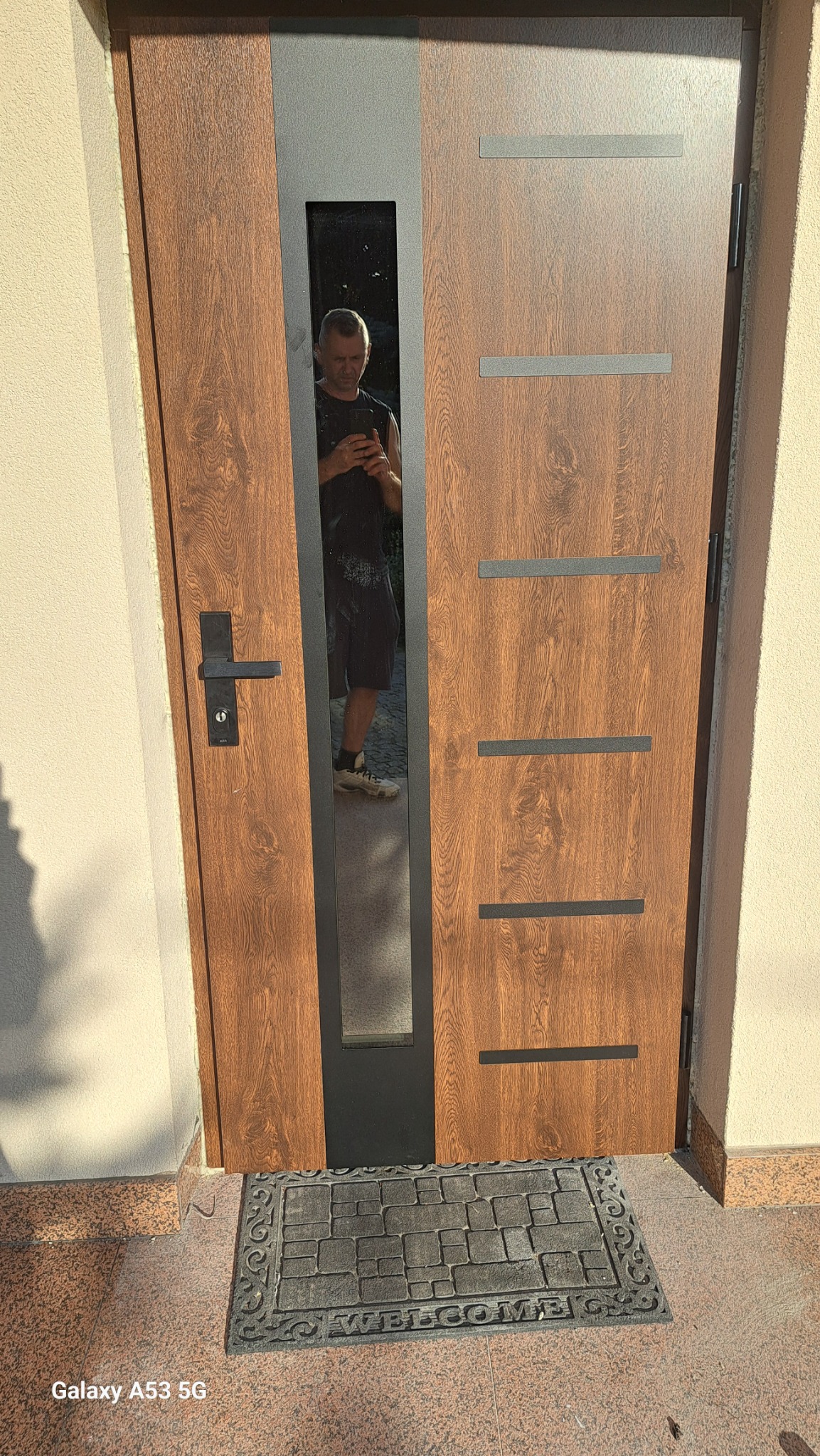 Brązowe drzwi zewnętrzne z szybą, w której odbija się osoba robiąca zdjęcie. Na podłodze wycieraczka 'Welcome'.