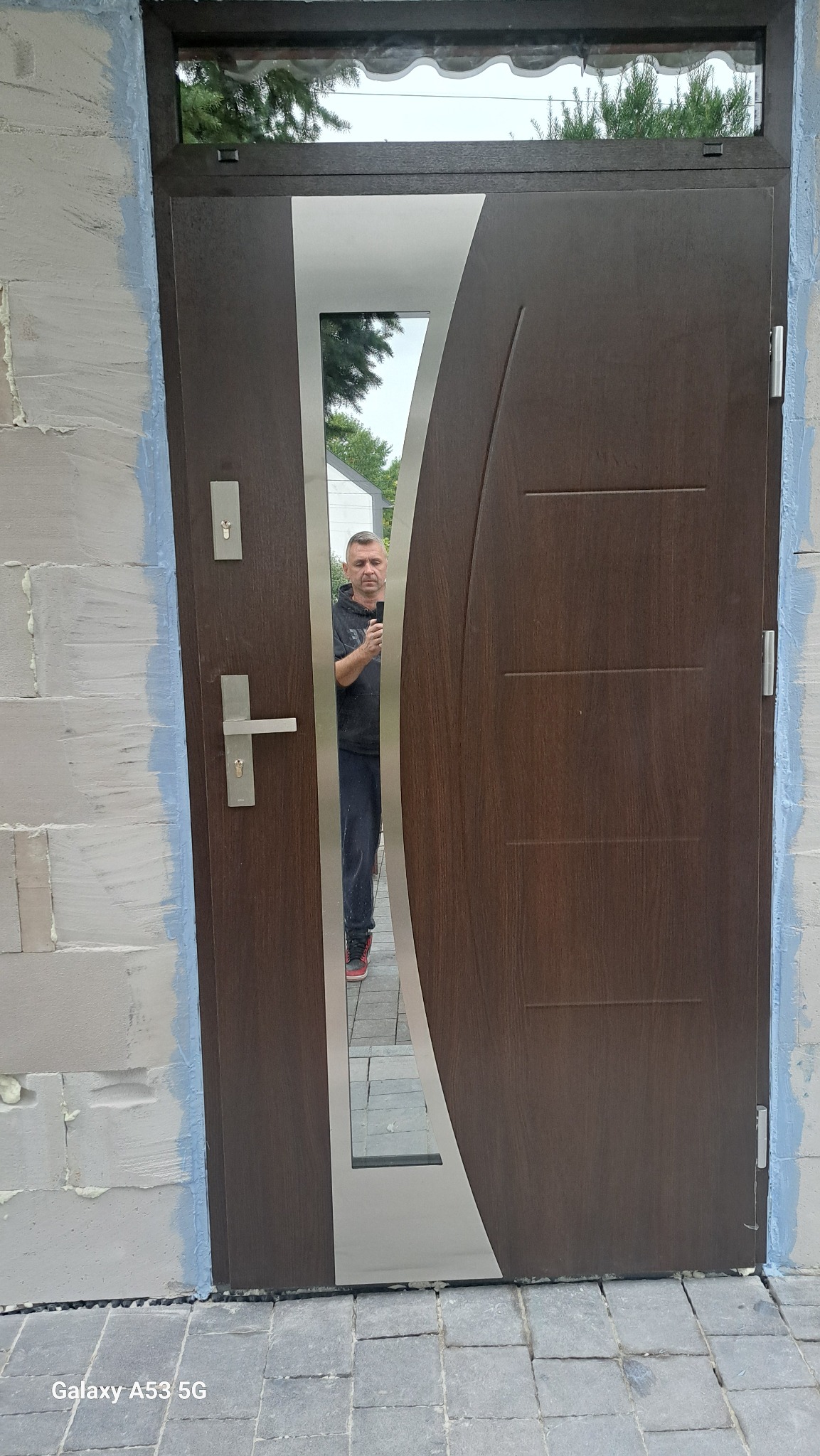 Nowoczesne drzwi zewnętrzne z przeszkleniem i brązowym wykończeniem, zamontowane w surowym murze z widocznym oknem nadprożowym. W lustrze odbija się osoba.