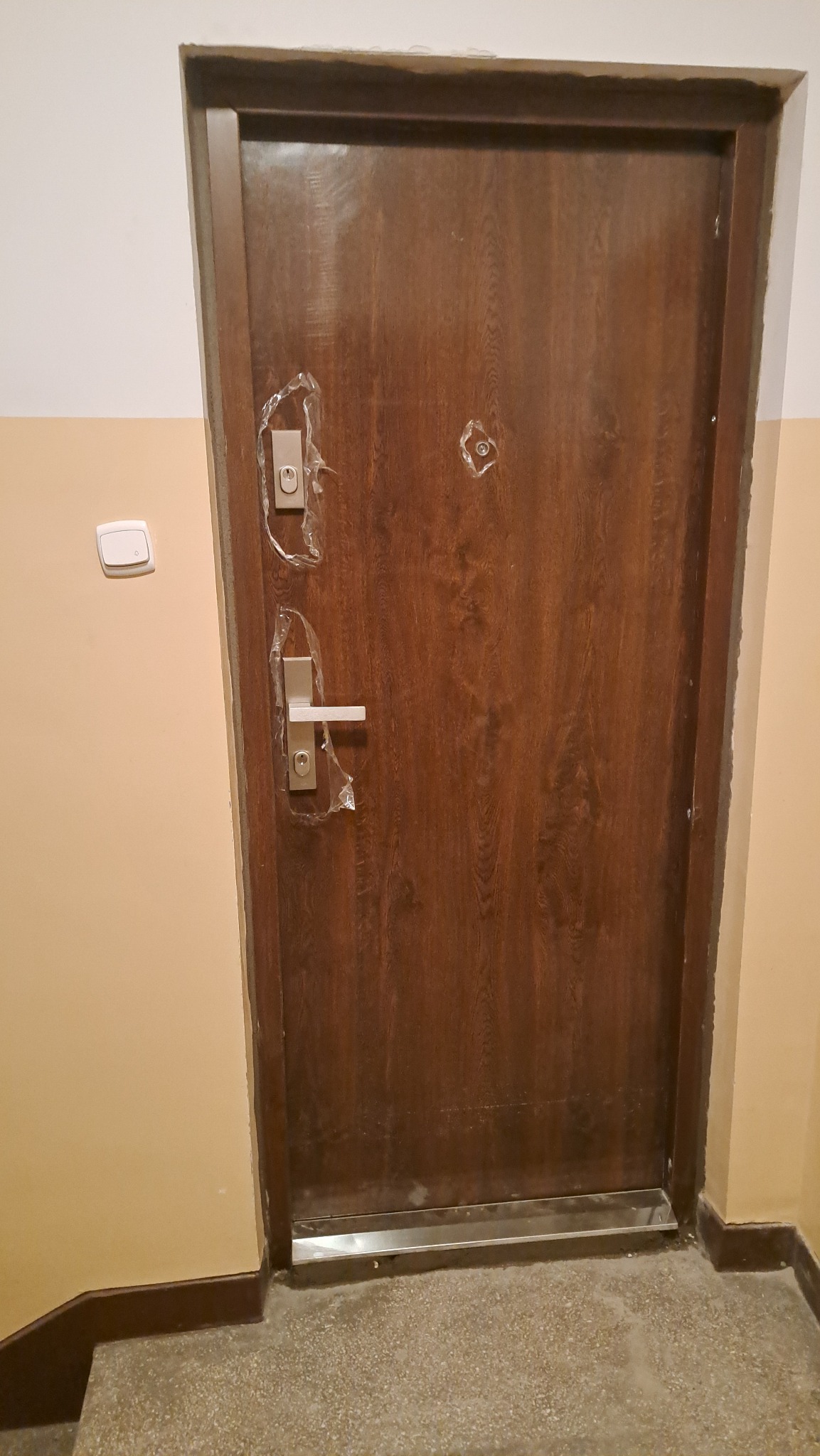 Ciemnobrązowe drzwi z metalową listwą progową i srebrną klamką, zabezpieczone folią ochronną, w korytarzu z beżowymi ścianami i szarym lastryko.