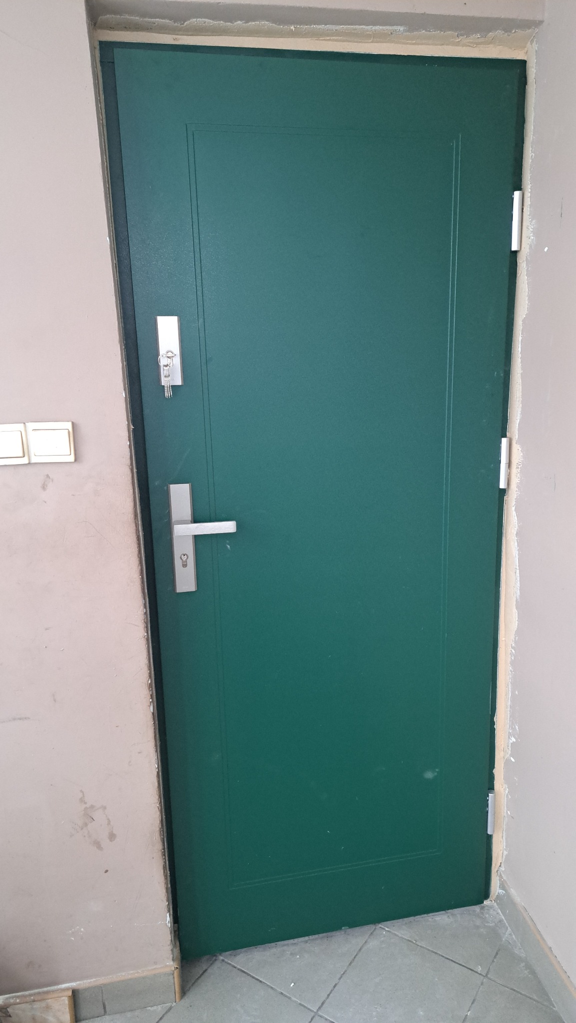 Zielone drzwi wejściowe z srebrną klamką i zamkiem, widoczne klucze w zamku, osadzone w surowym otworze drzwiowym, ściana w odcieniach beżu, płytki podłogowe.