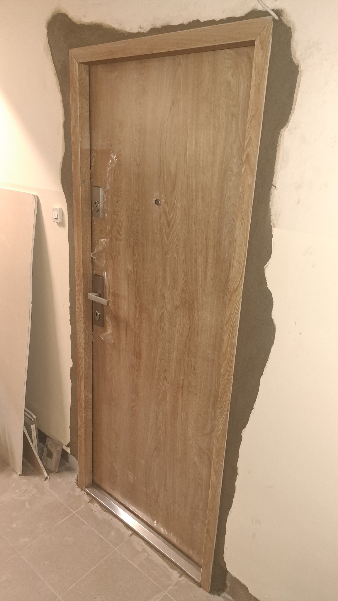 Jasne, drewnopodobne drzwi z metalową listwą progową, zamontowane w surowym otworze ściany, zabezpieczone folią. Widoczne ślady zaprawy i niedokończone wykończenie.