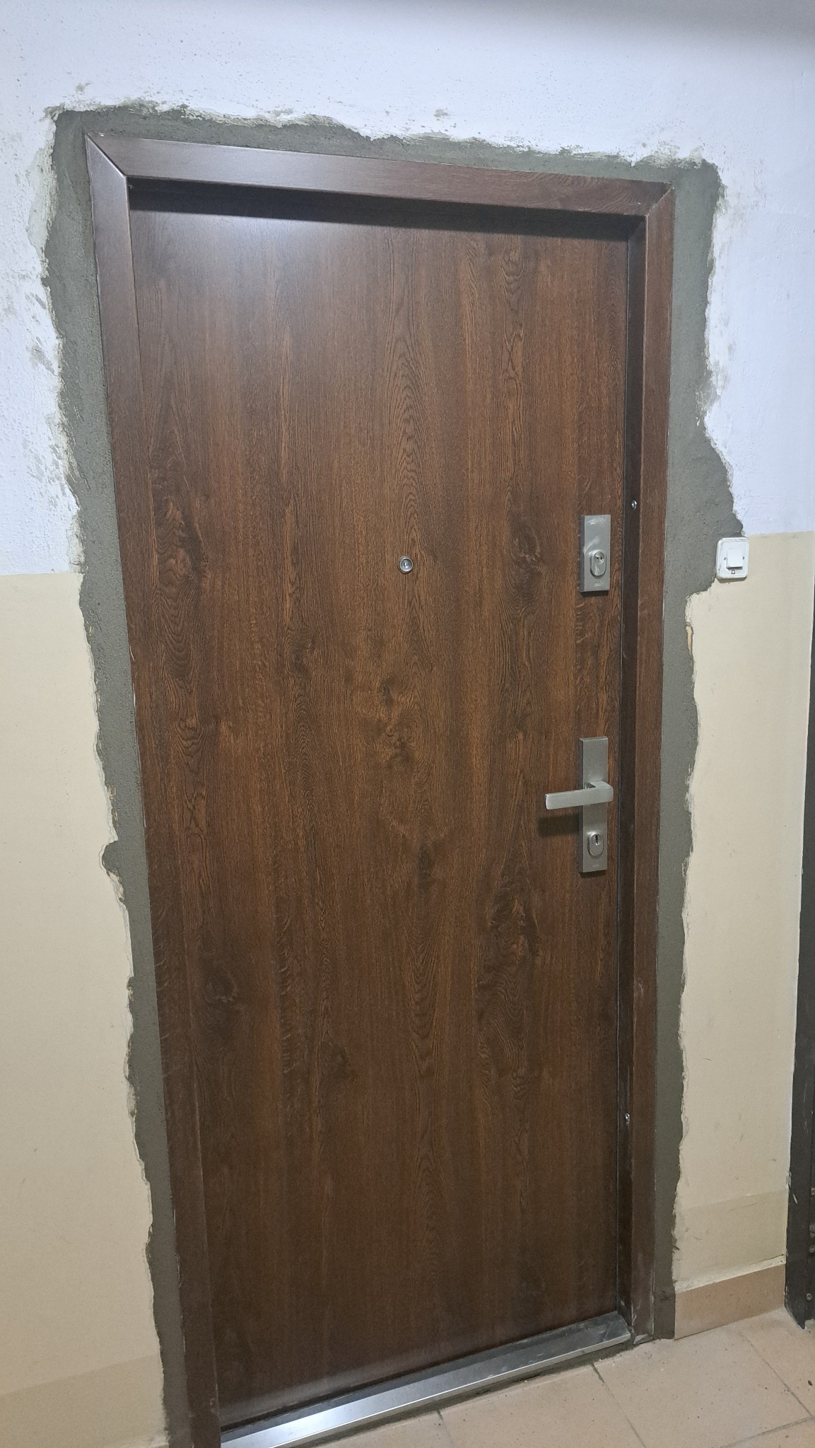 Brązowe drzwi z wizjerem i srebrną klamką, osadzone w surowym otworze z widocznym szarym cementem, ukazujące etap prac wykończeniowych.