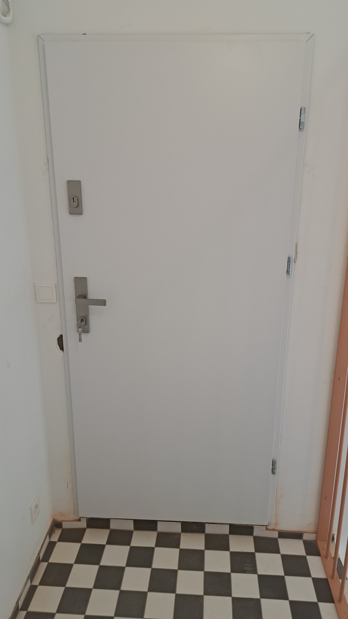 Białe drzwi z zamkiem i kluczem w korytarzu z czarno-białą podłogą w szachownicę. Widoczne zawiasy i listwa przypodłogowa.