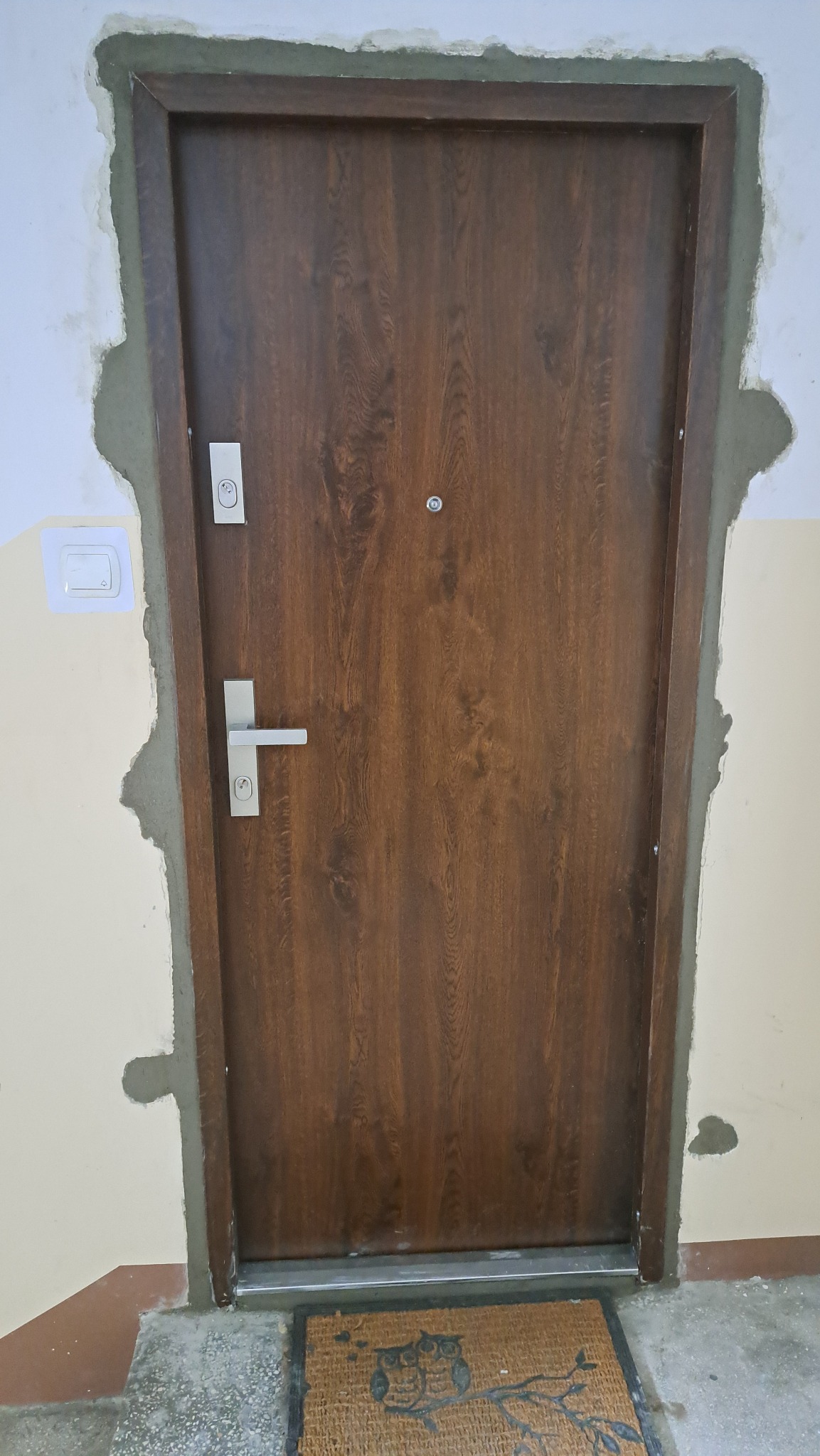 Ciemnobrązowe drzwi z wizjerem i srebrną klamką, osadzone w ościeżnicy z widocznymi śladami świeżego montażu i zaprawy. Wykończenie w trakcie prac, widoczna wycieraczka.