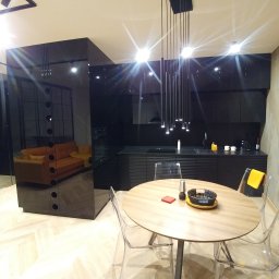 PROJEKT MG MARCIN GURTAT - Nowoczesna kuchnia w czerni z lakierowanymi frontami i subtelnym oświetleniem punktowym, widok na stół z przezroczystymi krzesłami i salon z sofami w odcieniach żółci i pomarańczy, oddzielone...