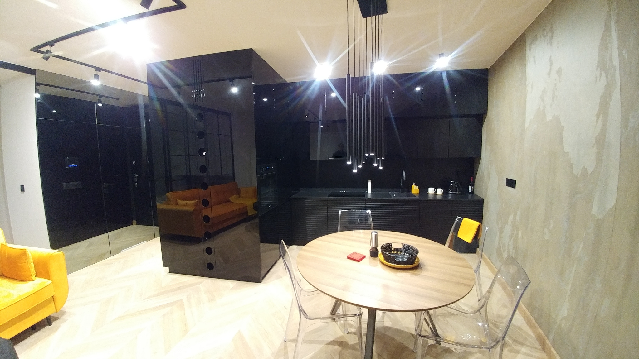 Nowoczesna kuchnia w czerni z lakierowanymi frontami i subtelnym oświetleniem punktowym, widok na stół z przezroczystymi krzesłami i salon z sofami w odcieniach żółci i pomarańczy, oddzielone...