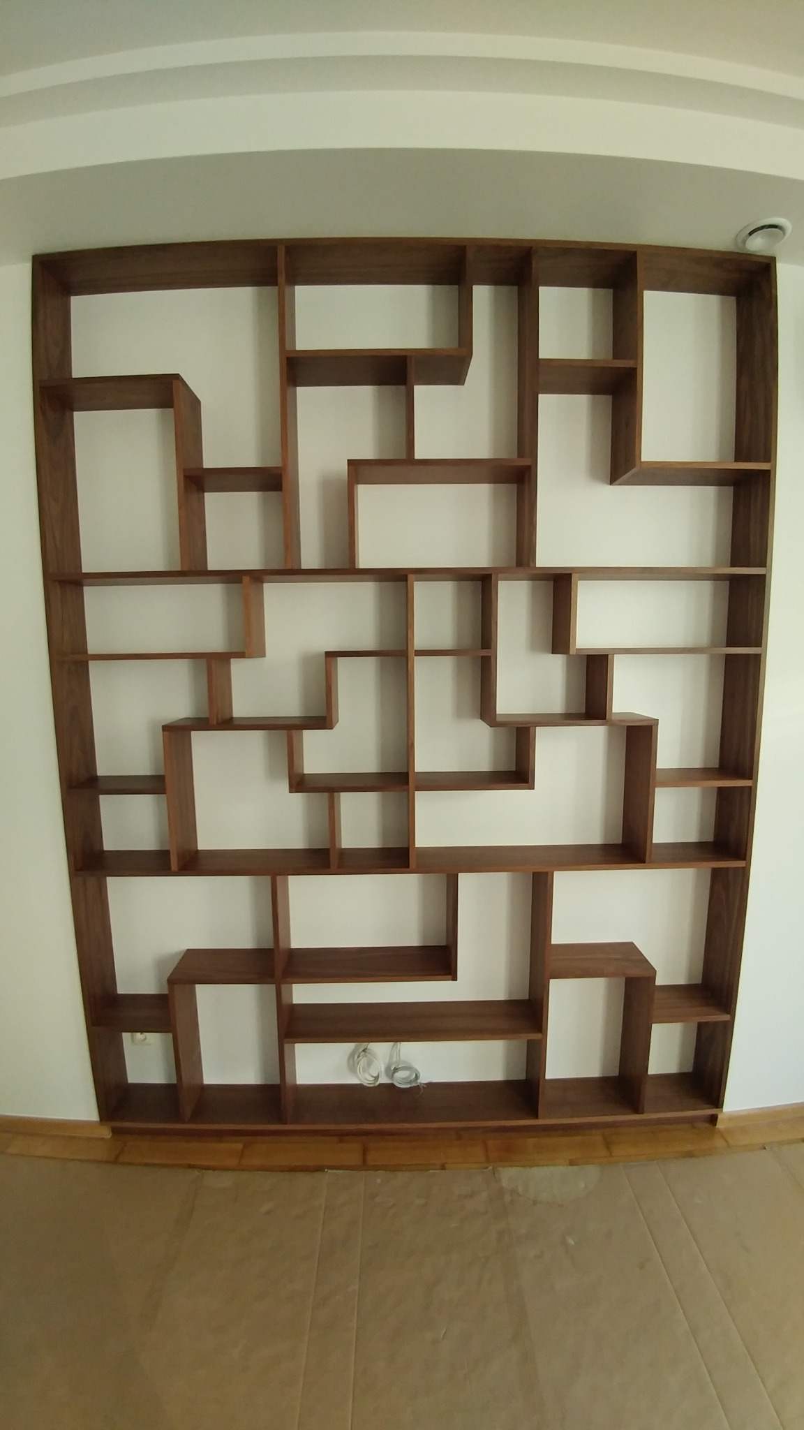 Fornirowany regał klejony o asymetrycznej konstrukcji z licznymi przegródkami i półkami, zamontowany na białej ścianie, z widocznymi przewodami elektrycznymi w dolnej części.