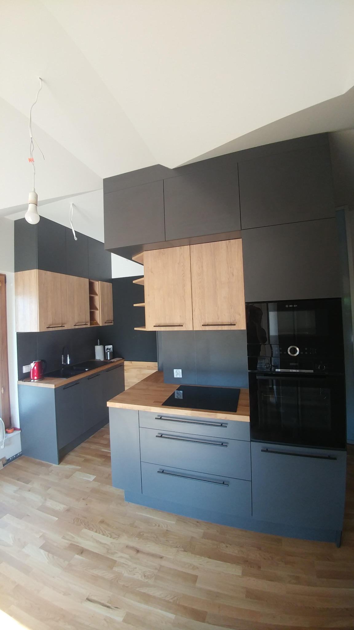 Nowoczesna kuchnia z szarymi dolnymi szafkami i drewnopodobnymi górnymi, drewniany blat, płyta indukcyjna i wbudowany piekarnik, surowa żarówka zwisająca z sufitu.