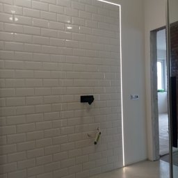 Usługi remontowo budowlane - Białe, prostokątne płytki na ścianie łazienki z oświetleniem LED wzdłuż krawędzi. Widoczne przyłącza wodne i fragment otworu drzwiowego.