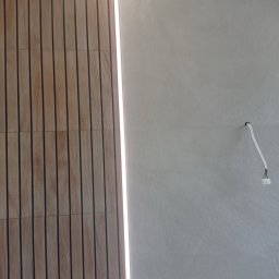 Usługi remontowo budowlane - Drewnopodobna okładzina ścienna z podświetleniem LED sąsiadująca z gładką, szarą ścianą z wystającym przewodem elektrycznym.