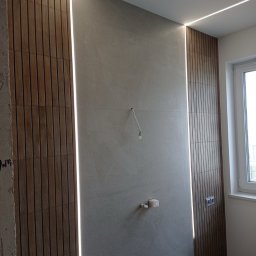 Usługi remontowo budowlane - Nowoczesna łazienka w trakcie wykańczania: drewnopodobna okładzina ścienna z podświetleniem LED, szara ściana, widoczne instalacje. Minimalistyczny design.