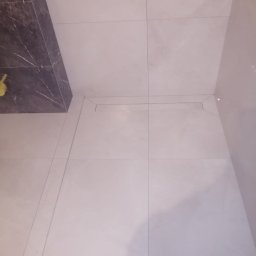 Usługi remontowo budowlane - Wykończona podłoga z jasnych, kwadratowych płytek w łazience w Wołominie. Widoczny odpływ liniowy i fragment ściany z ciemnego marmuru. Minimalistyczny design.