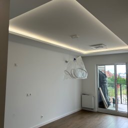Usługi remontowo budowlane - Nowoczesne wykończenie wnętrza: białe ściany, podwieszany sufit z oświetleniem LED, parkiet i zakryta lampa, w trakcie remontu, z widokiem na balkon.