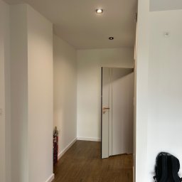 Usługi remontowo budowlane - Wnętrze korytarza z drewnianą podłogą, świeżo pomalowanymi, białymi ścianami i drzwiami z złotymi akcentami. Widoczny plecak na wiadrze farby.