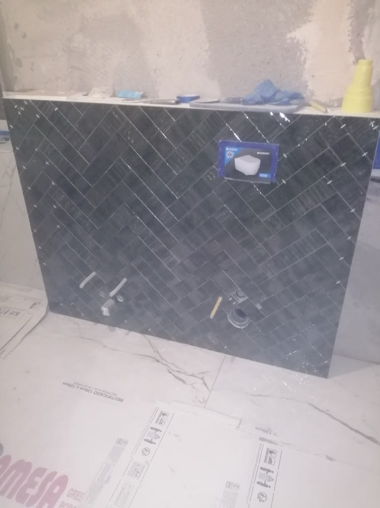 Obudowa stelaża WC wykończona ciemną, wzorzystą glazurą w jodełkę, z widocznymi przyłączami i pudełkiem po ceramice w tle, w trakcie prac wykończeniowych łazienki.