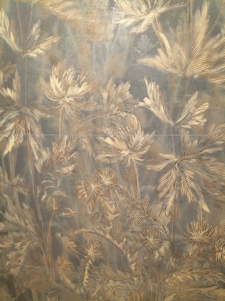 Położone płytki z motywem roślinnym w odcieniach beżu i szarości, tworzące dekoracyjną ścianę. Widoczne fugi.