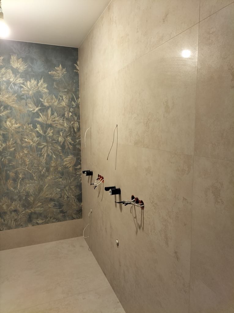 Łazienka w trakcie prac: beżowe płytki imitujące kamień, ściana z wzorem roślinnym, wyprowadzone kable elektryczne pod oświetlenie.