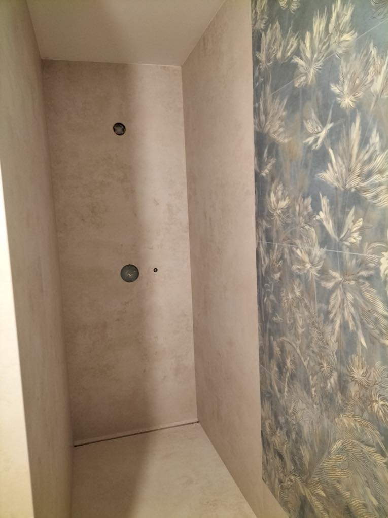 Wykończona kabina prysznicowa z beżowymi ścianami i dekoracyjną, niebieską ścianą z motywem roślinnym. Widoczne otwory na deszczownicę i baterię.