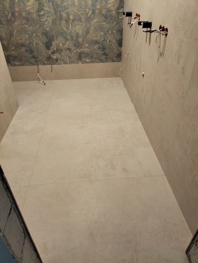 Wykończona łazienka z beżowymi płytkami na podłodze i ścianach, dekoracyjna tapeta na górze ściany, instalacja elektryczna przygotowana do montażu.